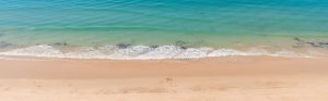 Imagen desde arriba de una playa con un barco a lo lejos, olas en la orilla, gente al sol y sombrillas de colores.